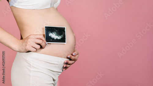 A happy expectant mother shows an ultrasound image of the fetus on camera. Prenatal ultrasound screening. close-up of the abdomen of a young pregnant woman holding a sonogram of an unborn child. A