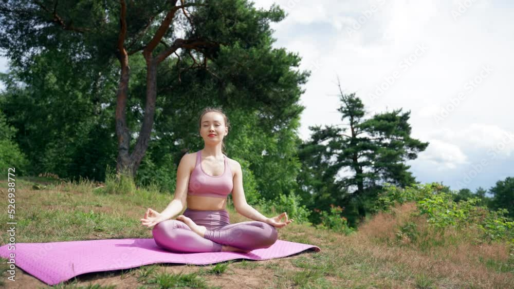 Young brunette meditates in Lotus pose with closed eyes on grassy meadow. Lady in pink sports suit sits smiling on mat against trees breathing fresh air Young brunette meditates in Lotus pose with closed eyes on grassy meadow. Lady in pink sports suit sits smiling on mat against trees breathing fresh air