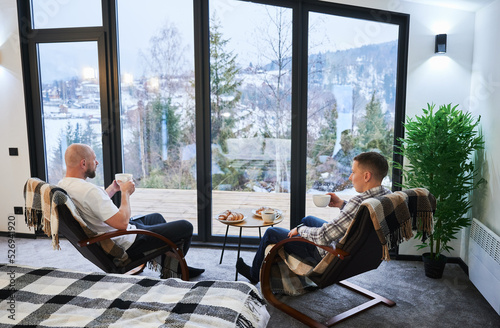 Two young men enjoying view of winter mountain landscape through panoramic windows in contemporary barn house. Happy male tourists sitting on chair, holding cup of tea in new cottage.