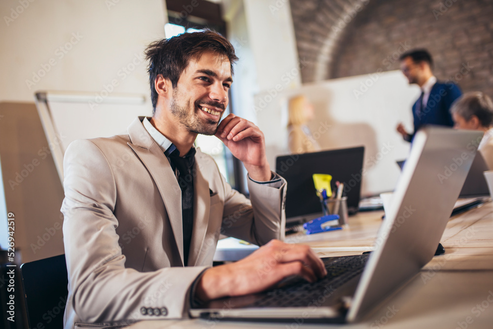 Happy businessman using laptop with team discussing project in the background.