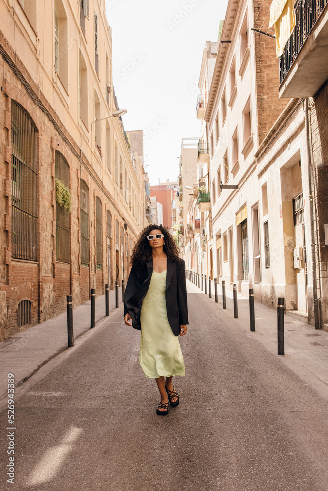 Fototapeta premium In full length young african woman walks alone on europe street in sunny weather. Curly brunette wears green coctail dress and black jacket. Concept vogue, stylish vacation.