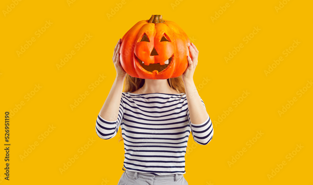Studio portrait of a woman with a pumpkin head. Funny young girl standing isolated on a yellow background, holding a carved orange pumpkin and hiding her face behind it. Halloween concept - obrazy, fototapety, plakaty 