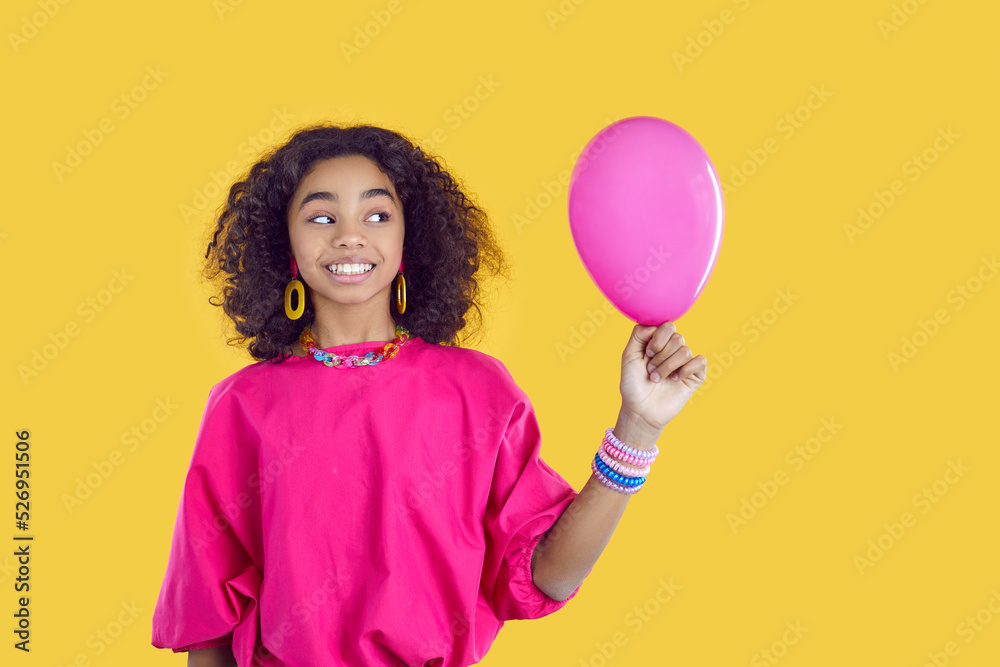 Cheerful beautiful dark-skinned teenage girl with funny expression looks at balloon in her hand. Happy curly girl in pink blouse and with colorful accessories who is preparing for birthday party. Cheerful beautiful dark-skinned teenage girl with funny expression looks at balloon in her hand. Happy curly girl in pink blouse and with colorful accessories who is preparing for birthday party.