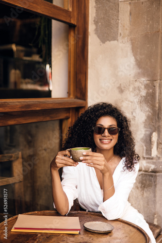 Nice young african girl in sunglasses looking at camera smiling, drinking coffee spring morning. Curly brunette hair woman wear casual clothes. Concept time to coffee. 
