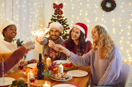 Smiling diverse friends with fireworks sit at table celebrate New Year together. Happy multiracial young people with sparkler lights enjoy Christmas party or celebration. Winter holidays.