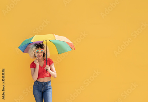 Smiling woman with prop rainbow and multi colored umbrella in front of yellow wall photo