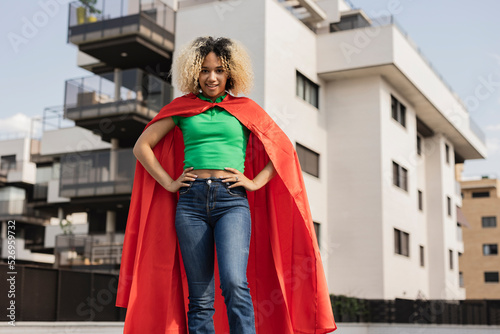 Smiling young woman wearing cape standing with hand on hip in front of wall photo