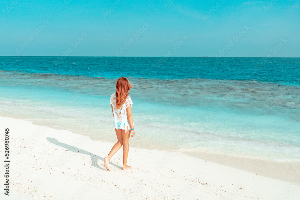 red haired cute teenage girl in swimsuit and cape stands on shore of the Indian Ocean in Maldives island, summer vacation