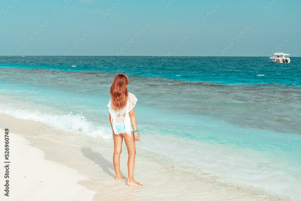 red haired cute teenage girl in swimsuit and cape stands on shore of the Indian Ocean in Maldives island, summer vacation