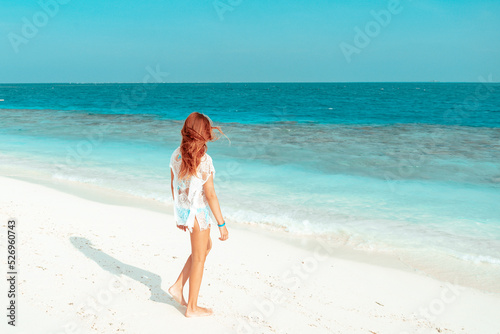 red haired cute teenage girl in swimsuit and cape stands on shore of the Indian Ocean in Maldives island  summer vacation