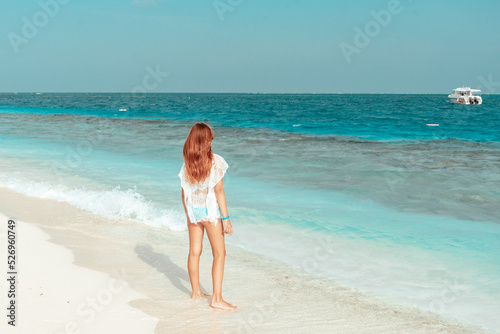 red haired cute teenage girl in swimsuit and cape stands on shore of the Indian Ocean in Maldives island, summer vacation