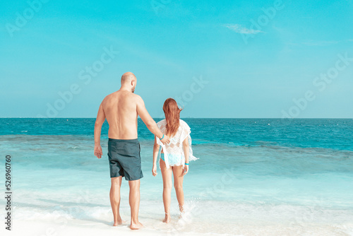 red haired cute teenage girl in swimsuit and young man father stand on shore of the Indian Ocean in Maldives island, family summer vacation