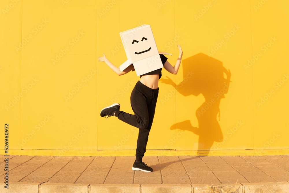 © VISTA by Westend61 - Playful woman wearing box with smiley face gesturing in front of yellow wall on footpath © VISTA by Westend61 - Playful woman wearing box with smiley face gesturing in front of yellow wall on footpath