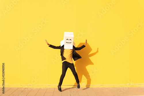 Happy man wearing box with smiley face jumping in front of yellow wall photo