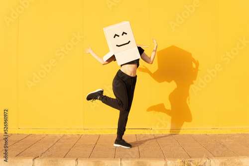 Playful woman wearing box with smiley face gesturing in front of yellow wall on footpath photo