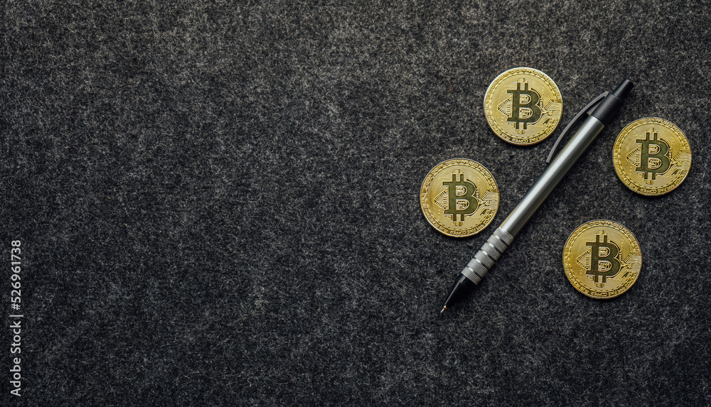 Gold bitcoin coin on dark gray felt desk pad with pen.