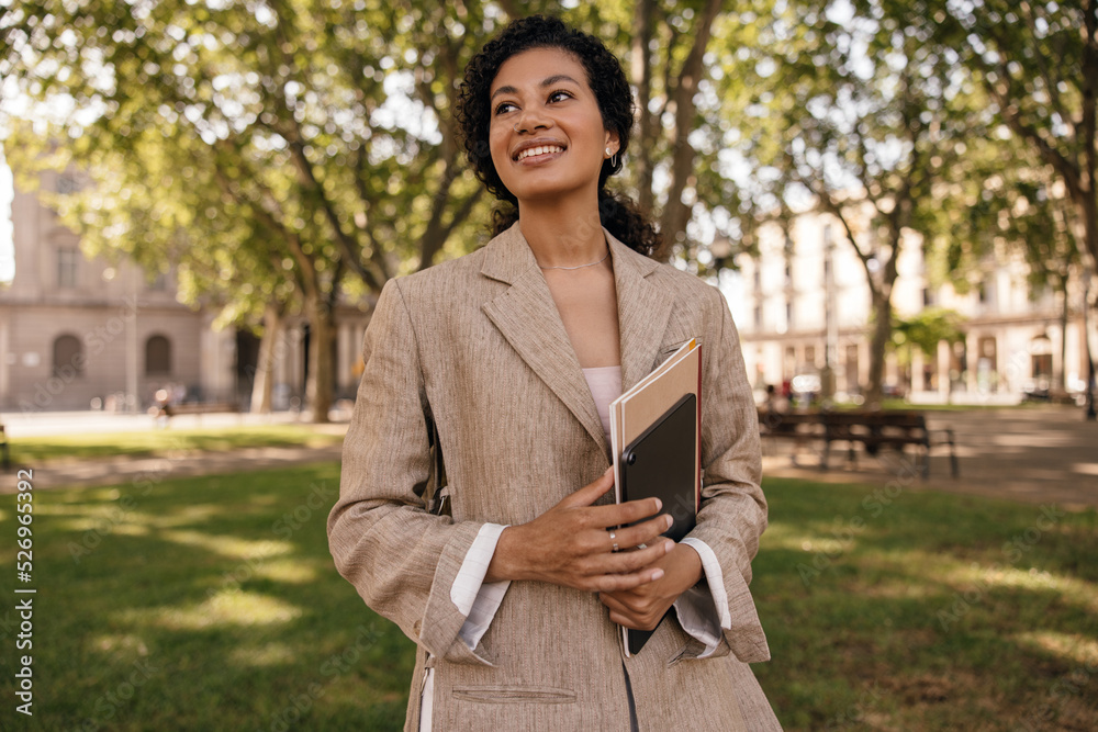Cheerful young caucasian woman looks away walks in park with folders and tablet. Brunette wears singlet and jacket. Concept student, good mood. Cheerful young caucasian woman looks away walks in park with folders and tablet. Brunette wears singlet and jacket. Concept student, good mood.