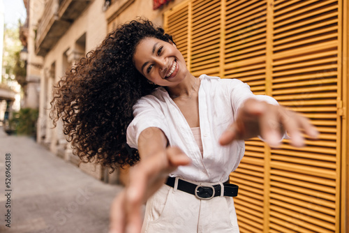 Happy young african woman looking at camera waving hair and stretches her hands in camera. Model with curly brunette hair wears singlet, shirt and white jeans. Concept city life.