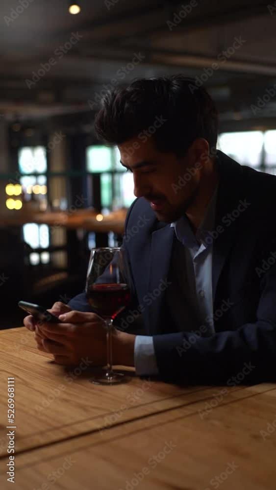 Vertical shot of confident young man in suit using smartphone during waiting for girlfriend, sitting at table with red wine in restaurant. Handsome bearded male waiting for female friend on meeting. Vertical shot of confident young man in suit using smartphone during waiting for girlfriend, sitting at table with red wine in restaurant. Handsome bearded male waiting for female friend on meeting.
