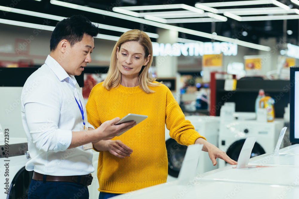 Obraz premium Asian consultant salesman in electronics and household appliances store, selling a working machine to a woman, recommending and approving the choice