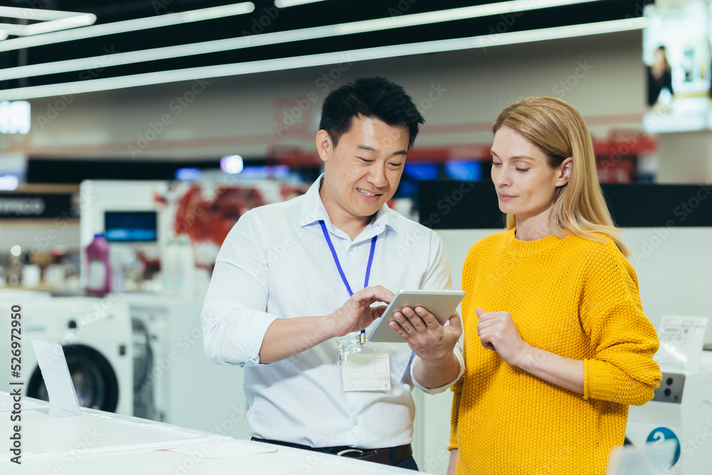 Obraz premium Asian consultant salesman in electronics and household appliances store, selling a working machine to a woman, recommending and approving the choice