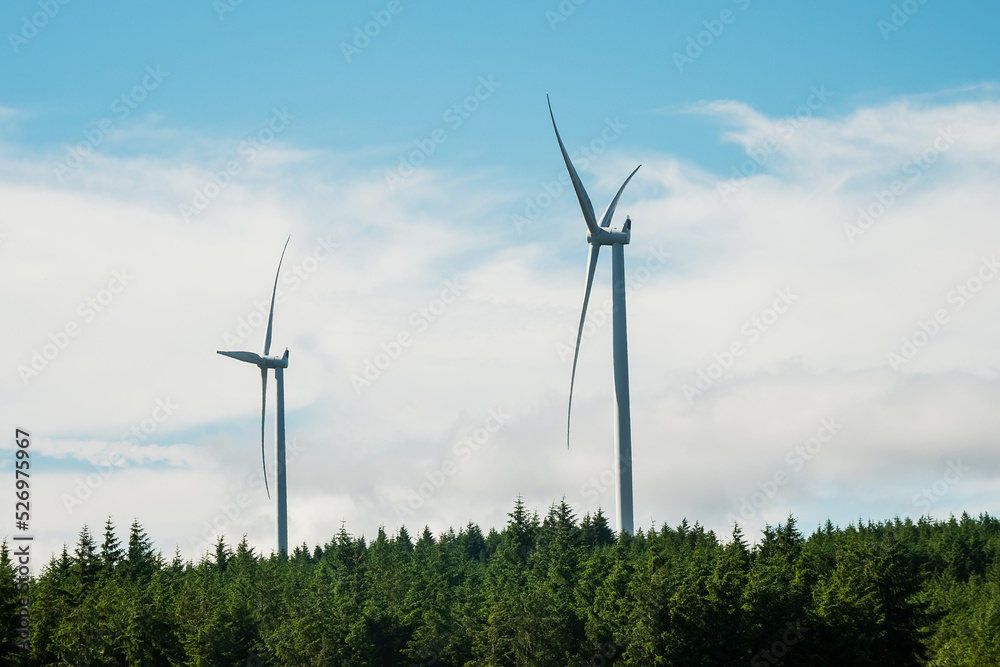 Wind electricity generator turbine in a forest area. Blending technology and nature environment. Production of clean renewable energy. Sunny day. Blue cloudy sky. Energy industry. Wind electricity generator turbine in a forest area. Blending technology and nature environment. Production of clean renewable energy. Sunny day. Blue cloudy sky. Energy industry.