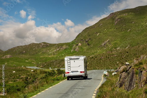Small motorhome on a small narrow asphalt road in a mountains. Wool sheep grazing grass on the road side. Warm sunny day, cloudy day. Connemara, Ireland. Travel and sightseeing.