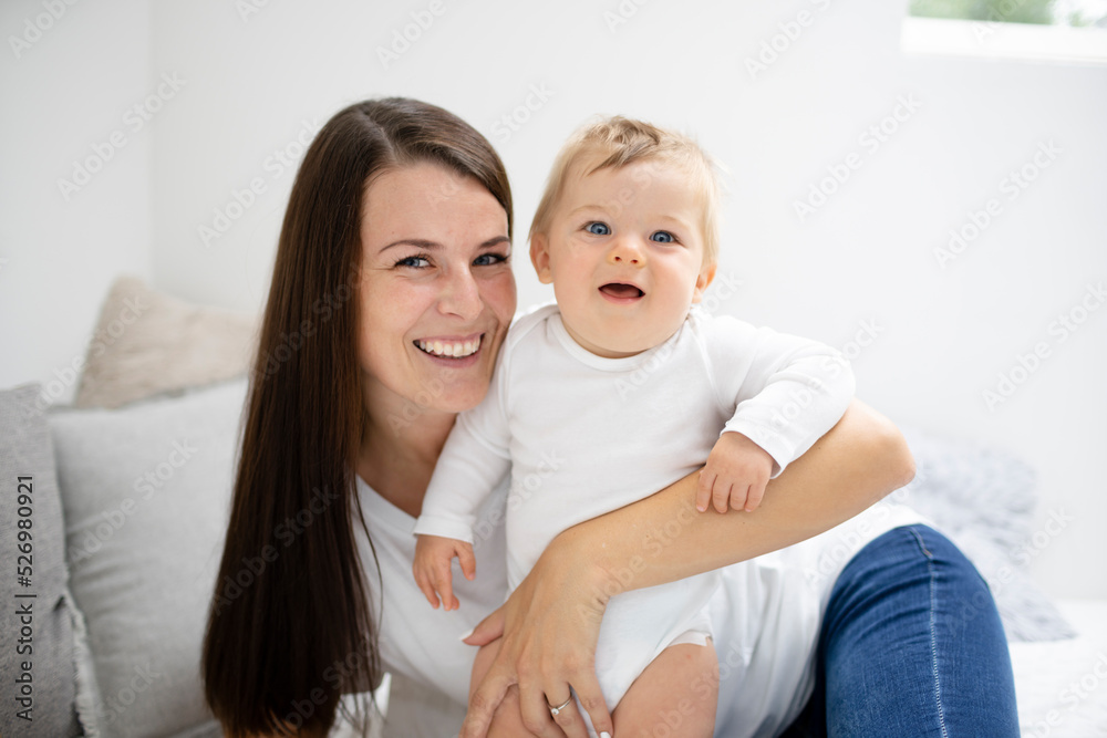 pretty young and happy mother with dark hair, white t-shirt, blue pants is cuddling in bed with her 7 months old son with blue eyes pretty young and happy mother with dark hair, white t-shirt, blue pants is cuddling in bed with her 7 months old son with blue eyes