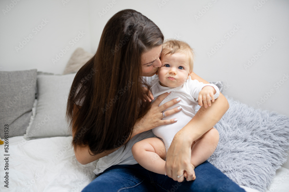 pretty young and happy mother with dark hair, white t-shirt, blue pants is cuddling in bed with her 7 months old son with blue eyes pretty young and happy mother with dark hair, white t-shirt, blue pants is cuddling in bed with her 7 months old son with blue eyes