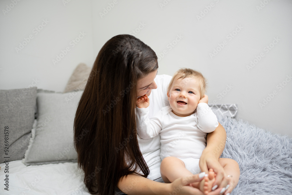 pretty young and happy mother with dark hair, white t-shirt, blue pants is cuddling in bed with her 7 months old son with blue eyes