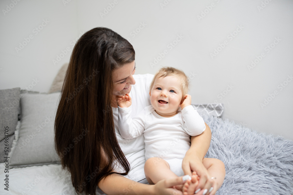 pretty young and happy mother with dark hair, white t-shirt, blue pants is cuddling in bed with her 7 months old son with blue eyes