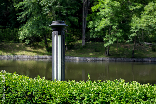 gray with white iron garden lantern of street lighting in boxwood bushes on background pond with water and park with trees, landscaping with lighting fixture object lit by sun summer day, nobody.