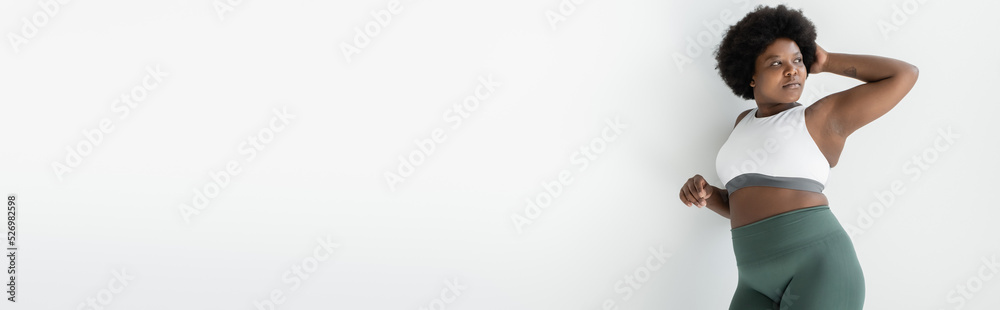 curly african american plus size woman in sportswear standing near white wall, banner.