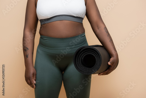 cropped view of tattooed african american plus size woman holding fitness mat isolated on beige.