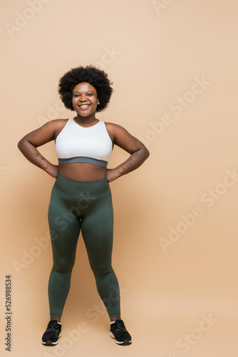 full length of happy african american plus size woman in crop top and leggings posing with hands on hips on beige.