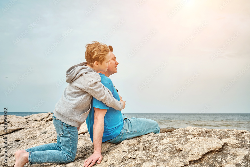 Happy family, father and son bonding, sitting on stone by the sea looking at view enjoying summer vacation. Togetherness Friendly concept
