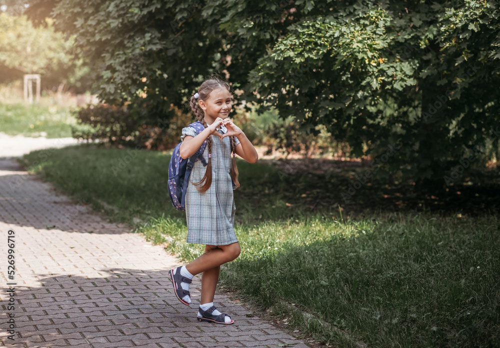 A little girl in a dress with pigtails holds large blue backpack and walks on road alley. First september day in elementary school.