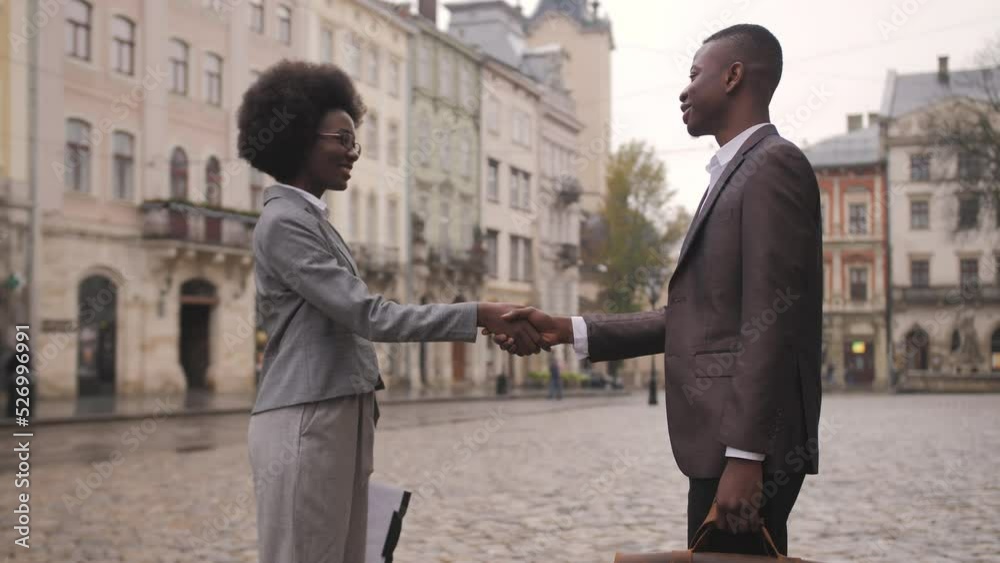 Two afro american partners in formal clothes shaking hands while standing on city street. Man holding leather briefcase, woman holding clipboard.