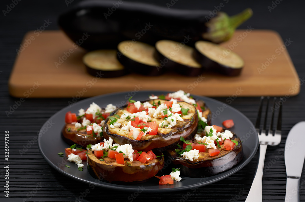 Grilled eggplant slices on a plate, with feta and tomatoes on a black table. whole eggplant background Grilled eggplant slices on a plate, with feta and tomatoes on a black table. whole eggplant background
