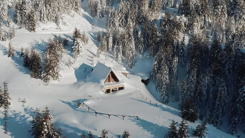 Drone shot of a small cottage house covered in snow on an icy winter morning on a sunny day. Winter fairytale in a remote forest.