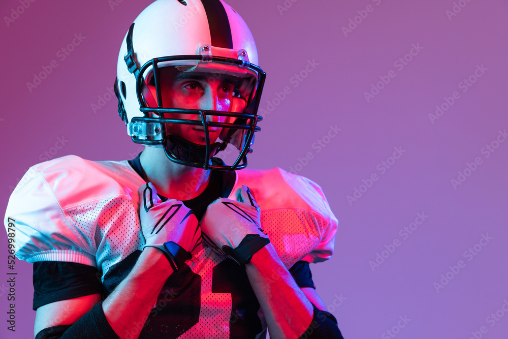 Close-up portrait of young man, american football player in helmet posing isolated over purple background neon light.