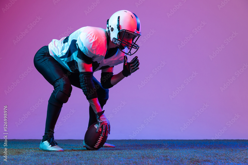 Portrait of concentrated man, professional american football player starting game, posing isolated over purple background in neon light.