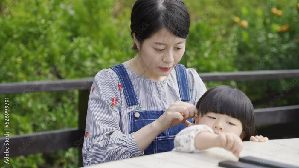portrait of a loving Asian mother doing up her innocent young daughter’s hair at an outdoor picnic table in the park on a sunny day.
