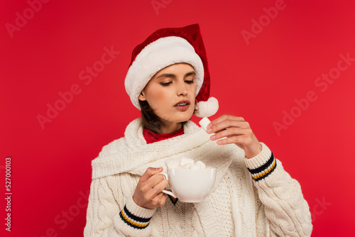 Young woman in santa hat holding cup and marshmallow isolated on red.