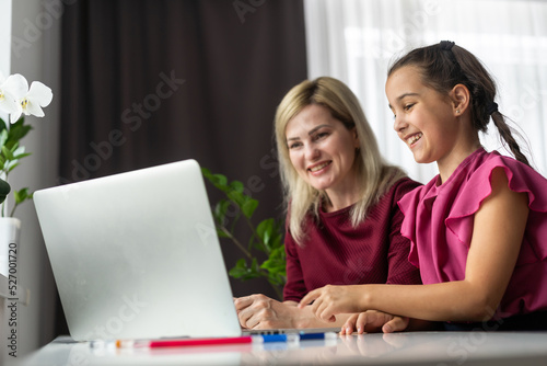 Pleased mother and small daughter sitting at table and school supplies and laptop and doing home task together in apartment