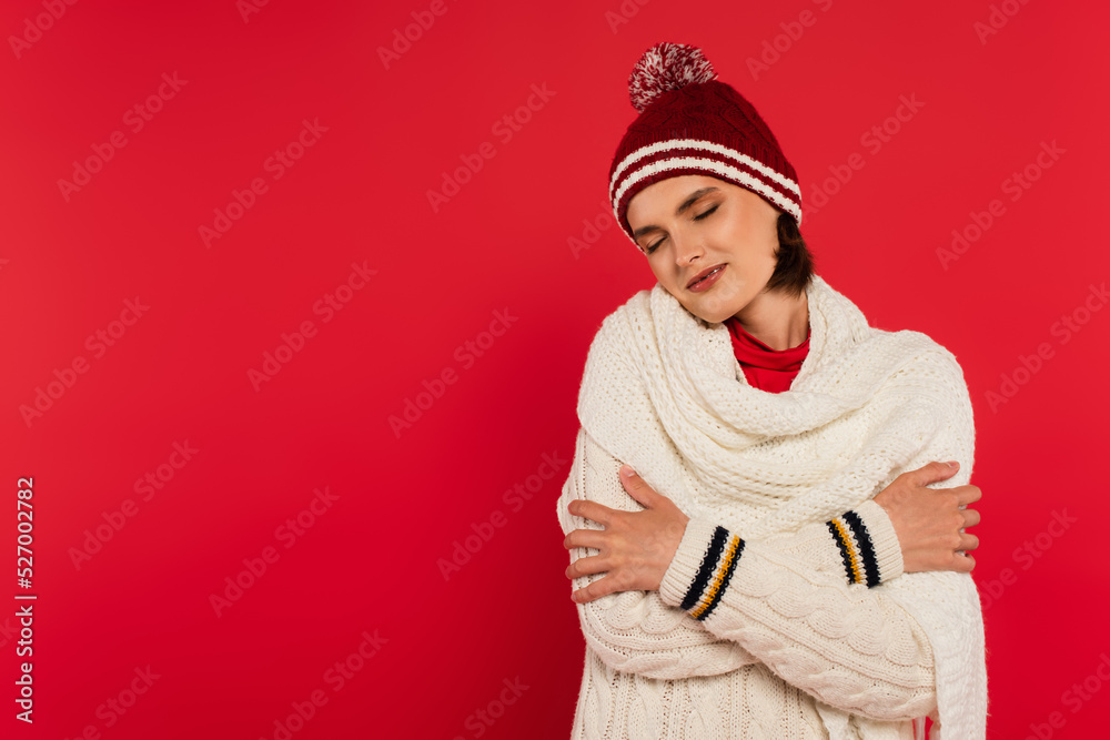Pleased woman in knitted clothes closing eyes isolated on red.