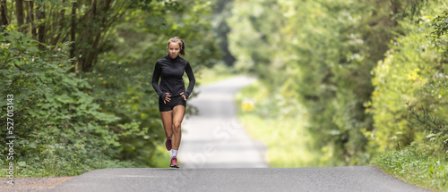 Fototapeta Naklejka Na Ścianę i Meble -  Young female athlete runs up the hill on an asphalt road in the nature