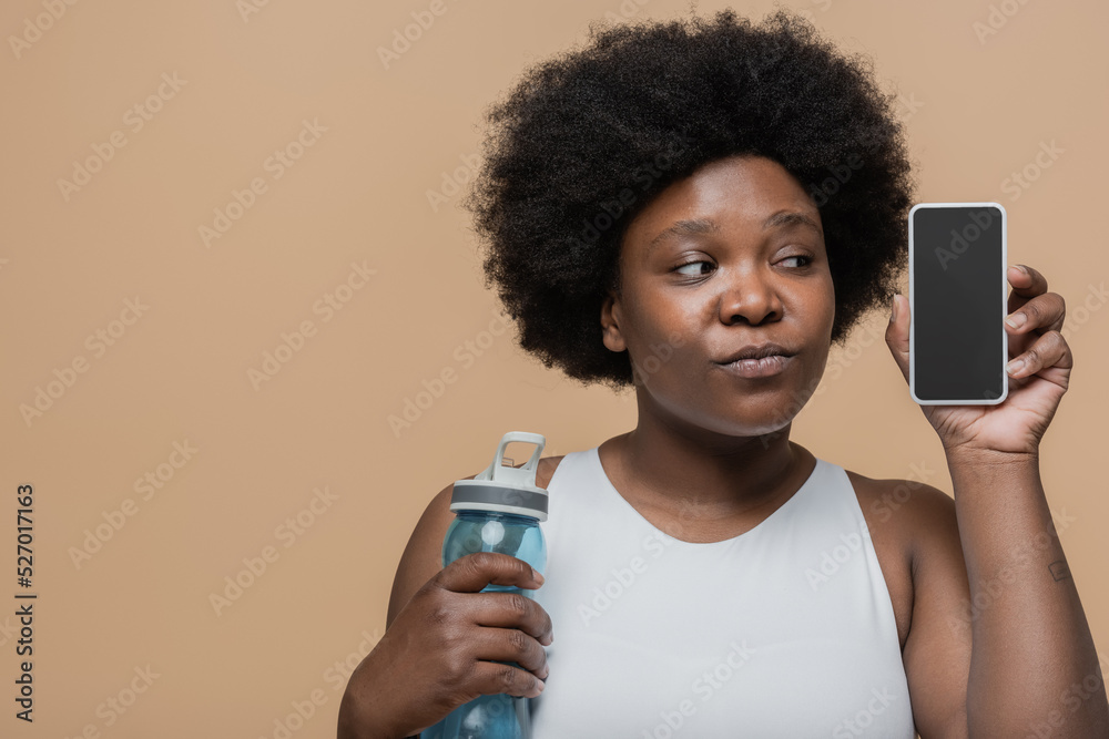 curious african american plus size woman holding sports bottle and smartphone with blank screen isolated on beige. curious african american plus size woman holding sports bottle and smartphone with blank screen isolated on beige.