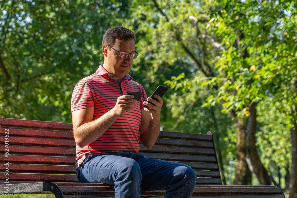 Obraz premium Happy millennial man using smartphone and credit card, sitting on wooden bench in sunny city park. Confident caucasian man purchasing online on cellphone
