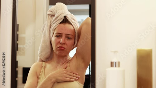 Unhappy young caucasian woman worried about black armpit with hair growing,looking in mirror reflection in bathroom at home.Feeling unsure with unwanted dark hair on the body, lacking self confidence